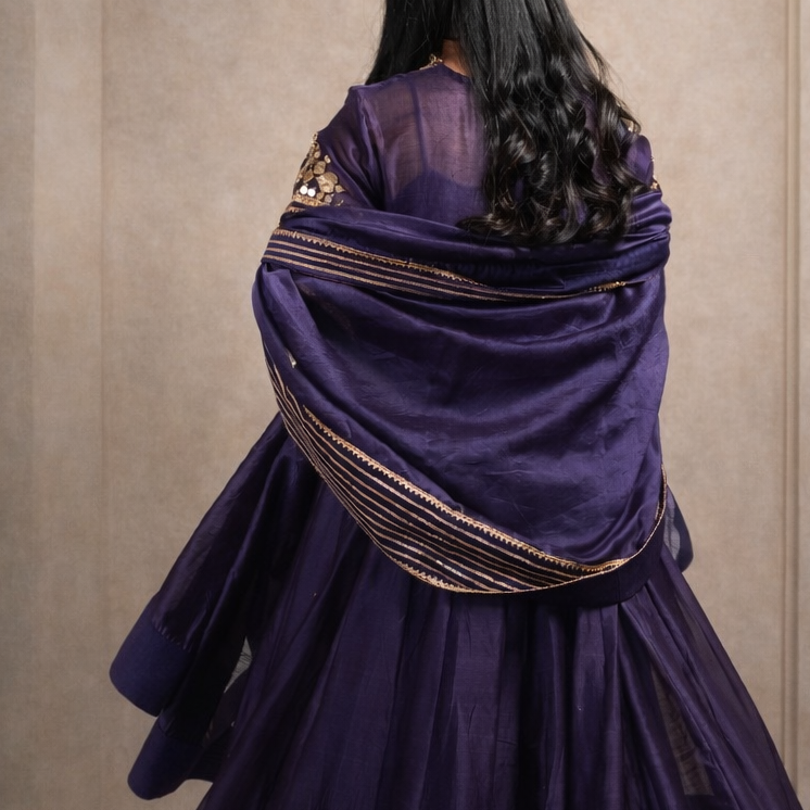 Person wearing a traditional purple outfit with gold accents in a neutral setting
